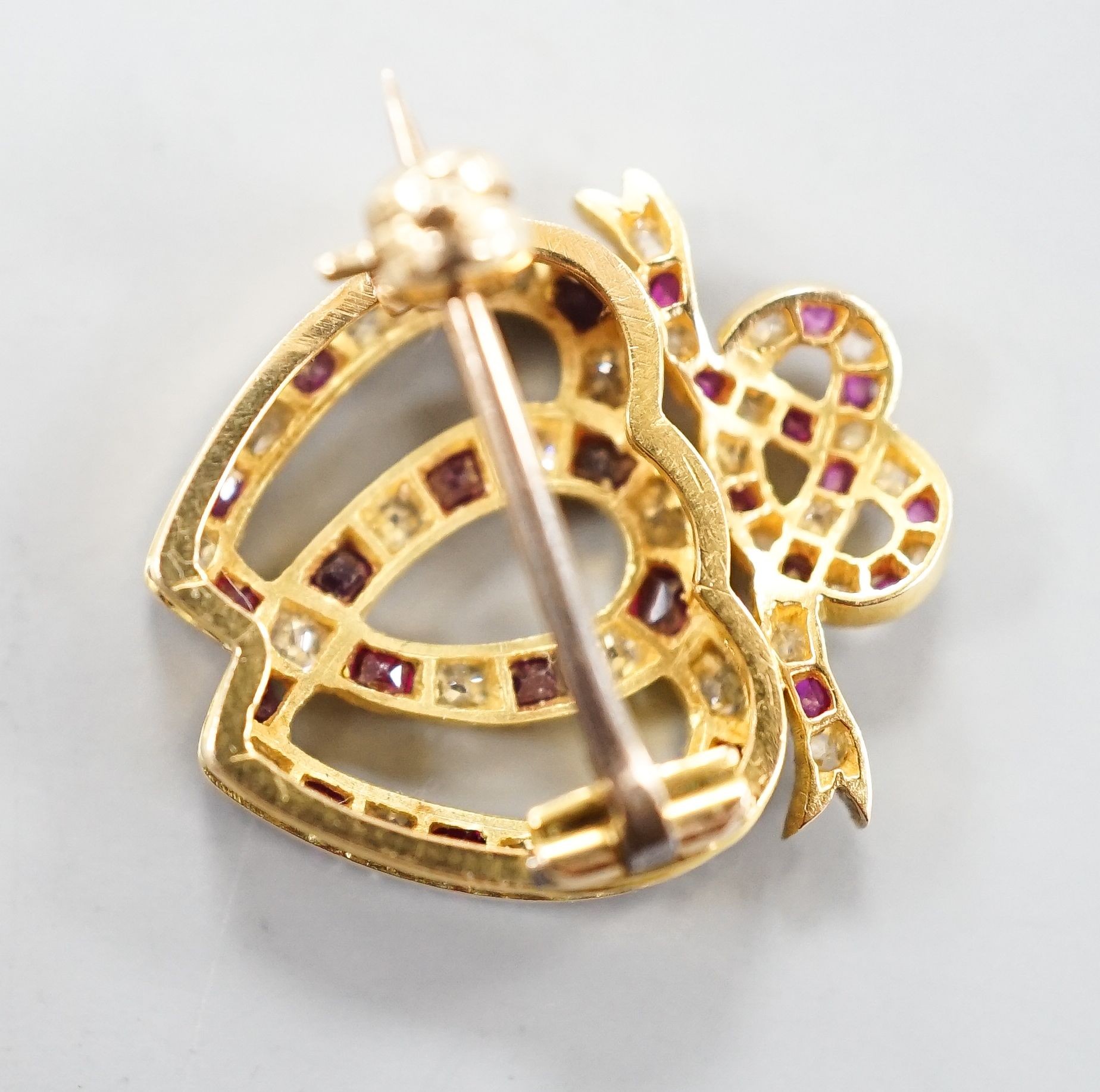 An early 20th century yellow metal, ruby and diamond set twin hearts brooch, with ribbon bow crest, 18mm, gross 2.4 grams.